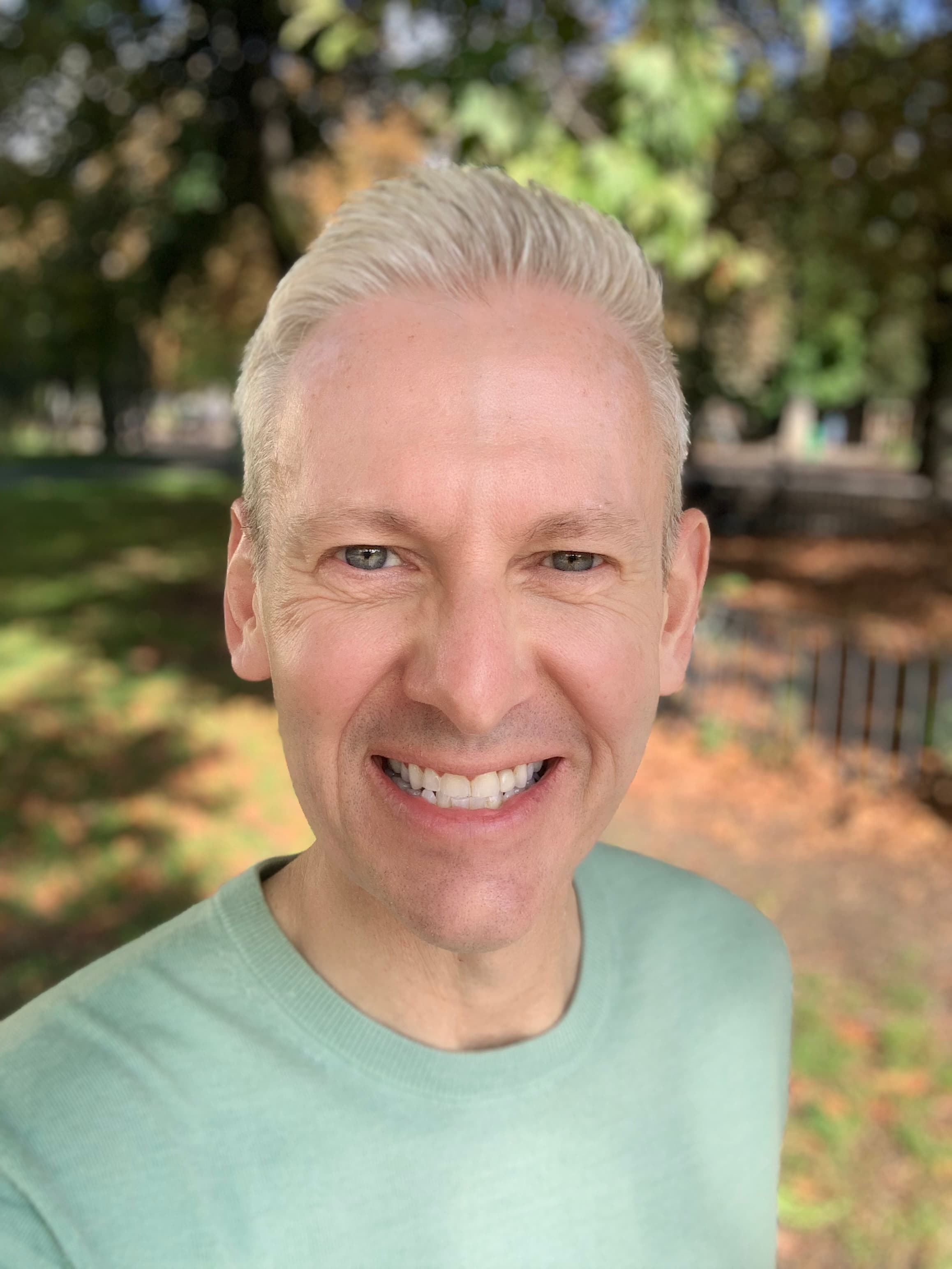 Smiling man with short blonde hair wearing a light green shirt in a sunny park.